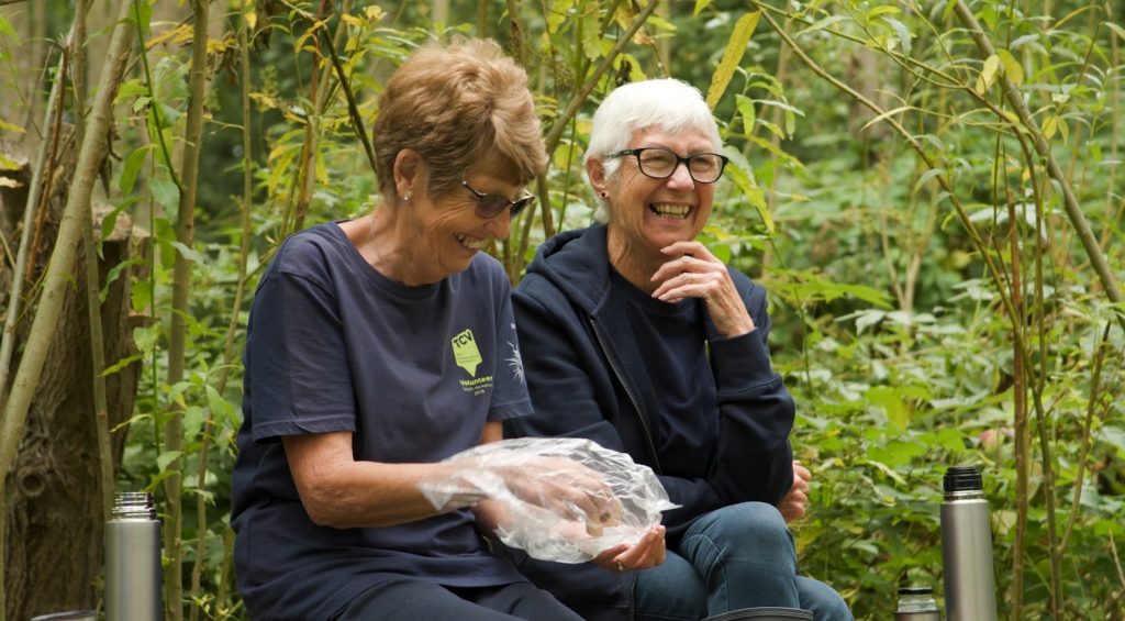 Two older ladies laughing while sat on a bench in woodland