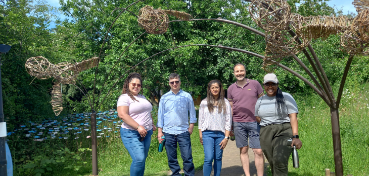 5 people stood outside under a large metal arch sculpture. They are all looking at the camera and smiling