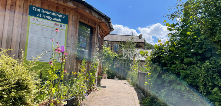 Image shows 'The Roundhouse' a round wooden building at TCV Hollybush. It is surrounded by plants and greenery, and there is a path running alongside the roundhouse.