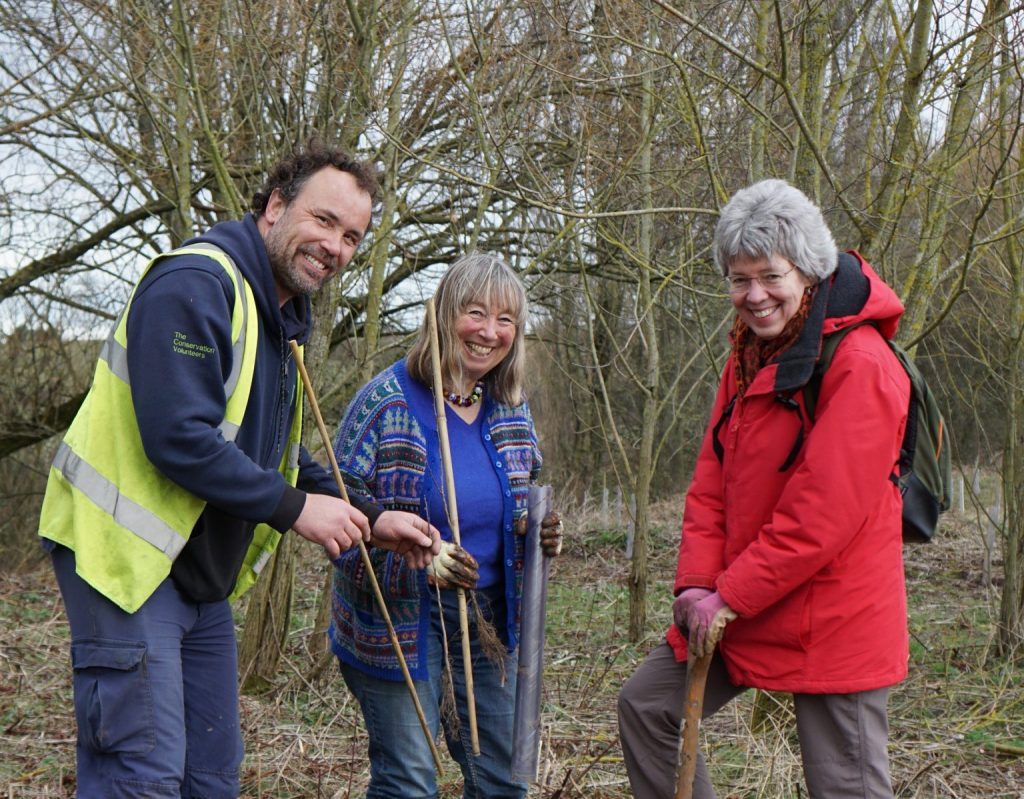 3 people smiling at the camera in woodland, holding trees ready to be planted