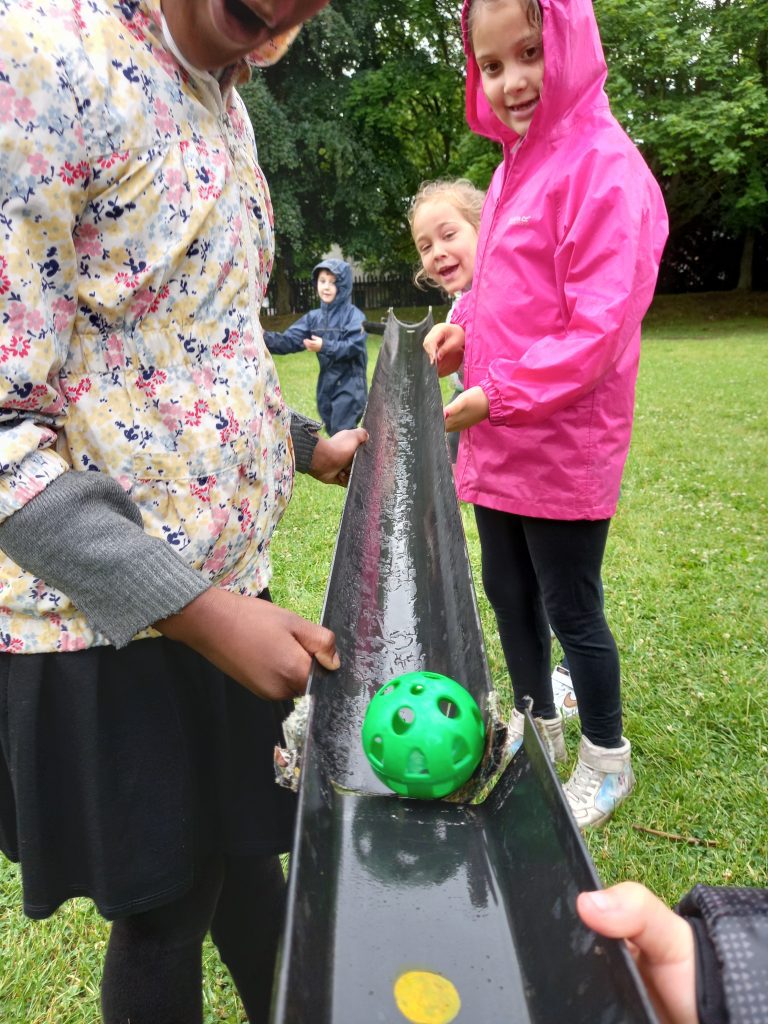 Photo shows a group of children rolling a green ball along a length of dark grey plastic guttering towards the camera. On the left is a girl wearing a multi-coloured jacket, and on the right is a girl wearing a pink jacket with her hood up and black leggings.
