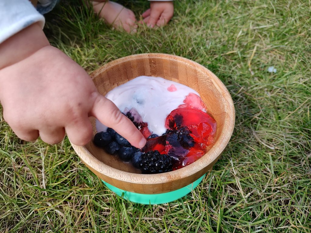 Bowl of berry and yoghurt pudding in a garden with a toddler's hand pointing to it.