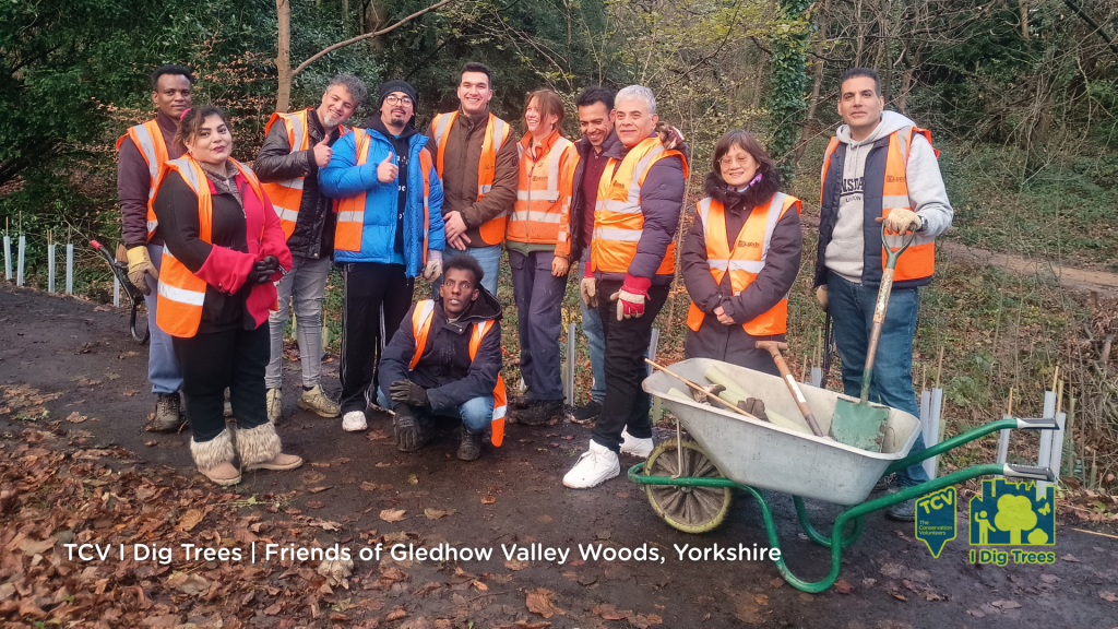 A cheerful group of people, in front of newly planted trees, in orange vests stands together for a photo, highlighting their unity and shared purpose, showcasing camaraderie and teamwork.