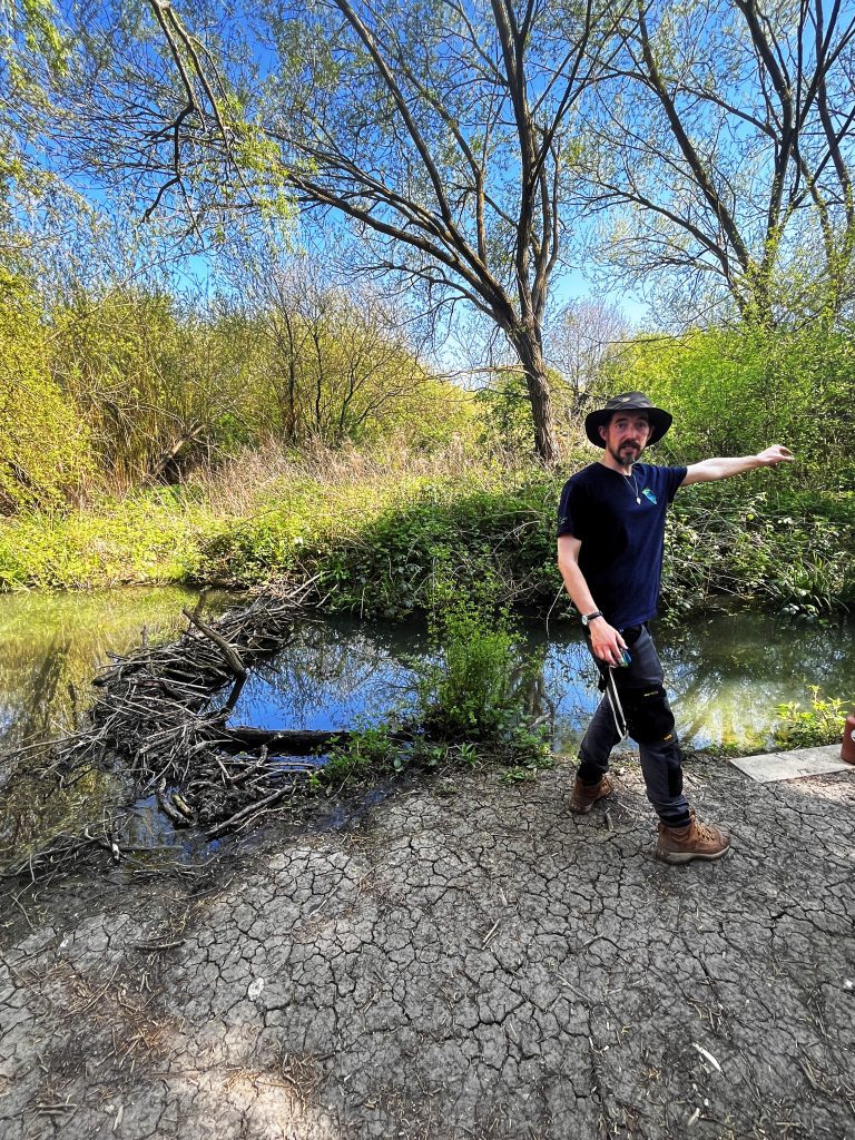 Man standing on a river bank with greenery and a blue sky background.