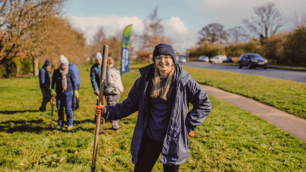 A smiling woman grips a rake while standing on a grass verge, preparing for wildlife-friendly gardening work.